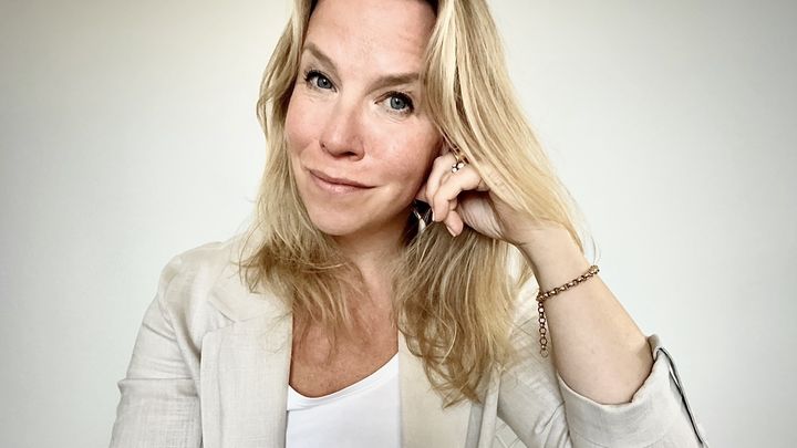 Vaaleaan jakkuun pukeutunut vaaleahiuksinen nainen katsoo kameraan.