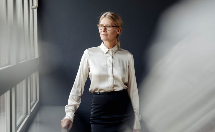 Nainen seisoo ikkunan ääressä katsellen sivulle. Hänellä on yllään valkoinen paita ja musta hame.