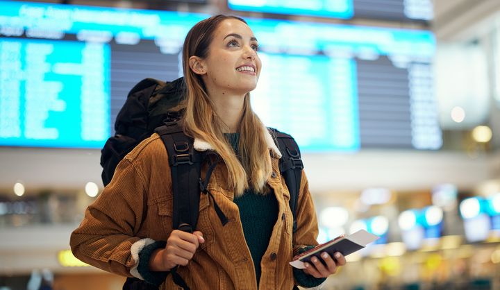 Nainen katsoo ylöspäin lentokentällä, kantaa reppua ja pitää kädessään passia ja lippuja.