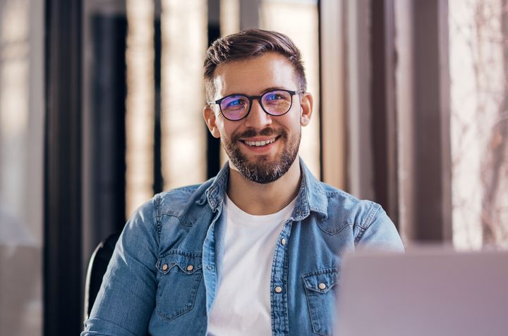 Mies istuu tietokoneen ääressä ja hymyilee kameralle. Hänellä on silmälasit ja päällään farkkupaita.