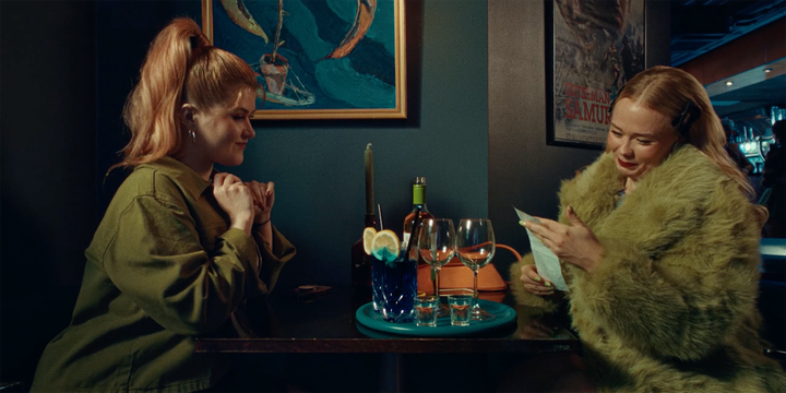 Kaksi naista istuu baarissa pöydän ääressä juomien kanssa. Toinen lukee listaa tai menua.