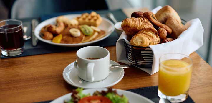 Aamiaispöytä, jossa on kahvikuppi, appelsiinimehu, croissantteja, sämpylöitä, hedelmiä ja leivonnaisia.