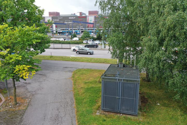 Harmaa ilmanlaadun mittausasema seisoo nurmikolla ja taustalla näkyy paljon autoja ja Sello-kauppakeskus.