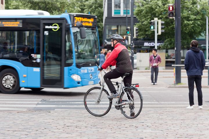 Linja-auto ajaa suojatien ohi, jossa pyöräilijä ja jalankulkijat odotttavat liikennevalon vaihtumista vihreäksi.