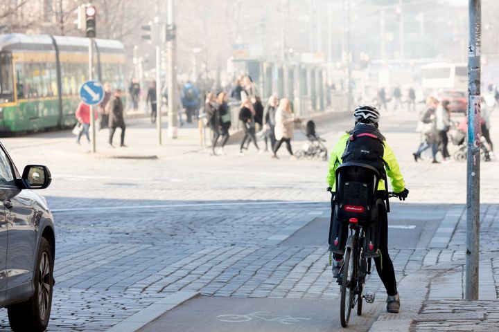 Pyöräilijä seisoo liikennevaloissa ja ympärillä näkyy jalankulkijoiden kulkevan sankan katupölypilven seassa.