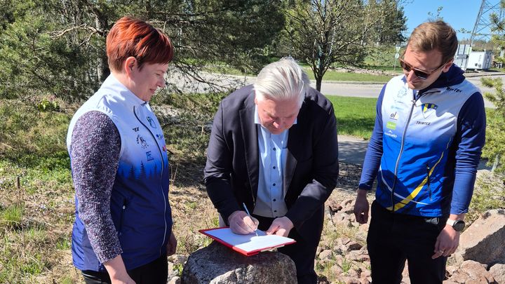 Kuvassa kolme henkilöä ulkona allekirjoittavat sopimuksen . Keskellä oleva henkilö kirjoittaa nimensä sopimukseen.