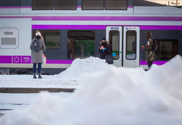 Kuvassa juna asemalla, asemalaiturilla seisoo kolme ihmistä lumikasojen keskellä