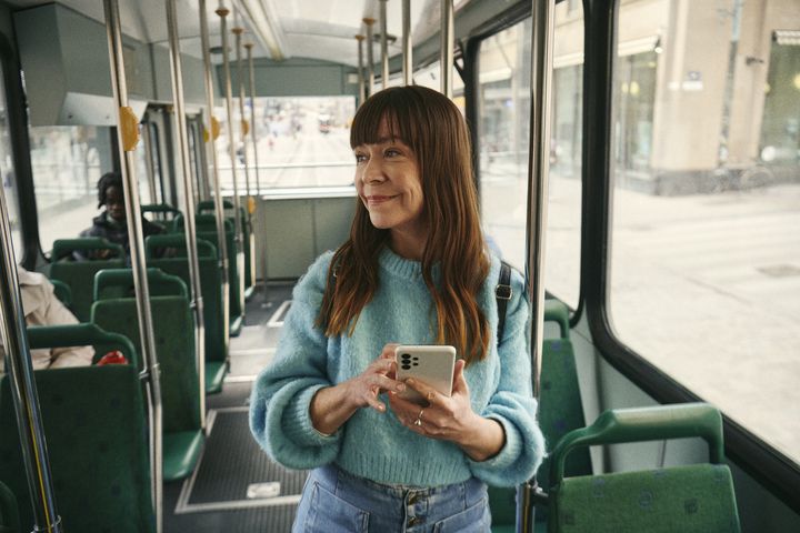 Nainen seisoo raitiovaunussa iloisena puhelin kädessään ja katsoo ulospäin