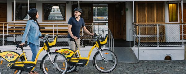 Kaksi kaupunkipyöräilijää saeisahtuneena lautan eteen kesäisessä maisemassa,