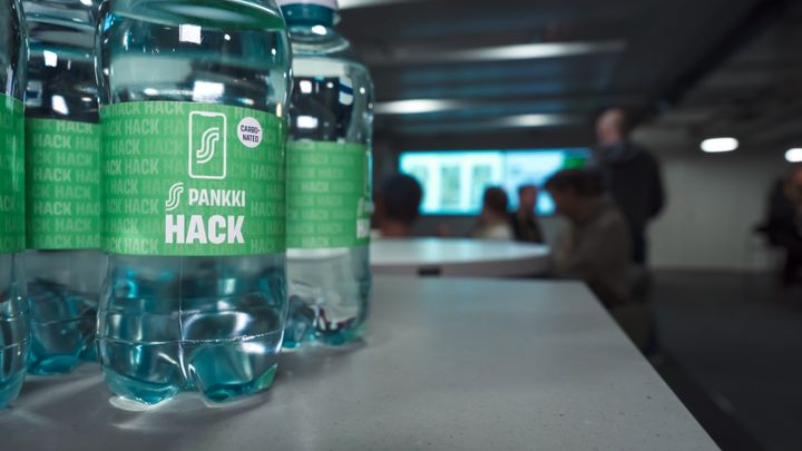 Läpinäkyviä vesipulloja, joissa on "S-Pankki Hack" -etiketti, pöydällä. Taustalla näkyy ihmisiä ja epätarkka näyttö.