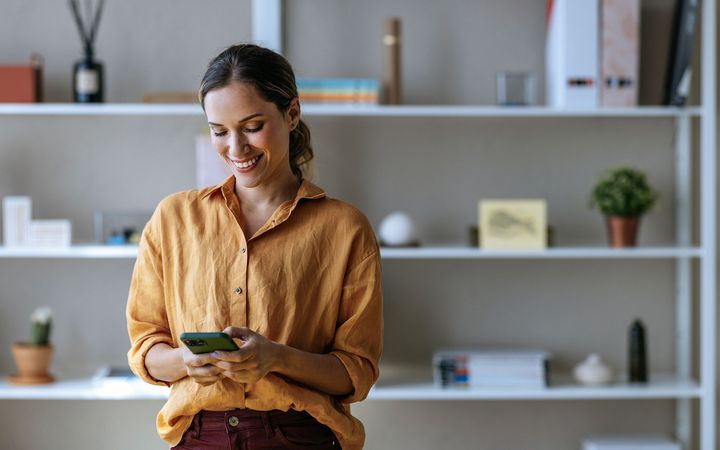 Kvinna står leende framför en bokhylla och tittar på sin mobiltelefon.