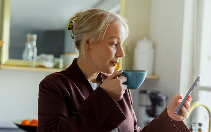 En person står i köket med en kaffekopp i handen och tittar på sin smarttelefon. I bakgrunden syns ett hemmakök i ljusa färger.