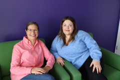 Kaksi naista istuu vierekkäin vihreillä tuoleilla ja hymyilee kameralle. Tausta on violetti.