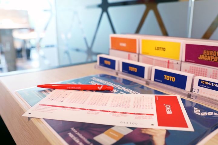 Vihkolomake ja punainen kynä pöydällä, taustalla lotto- ja arpalippuja telineessä.