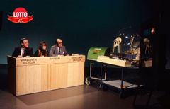 Kolme henkilöä istuu lottostudiossa vuonna 1981, seuraten arvontaa.