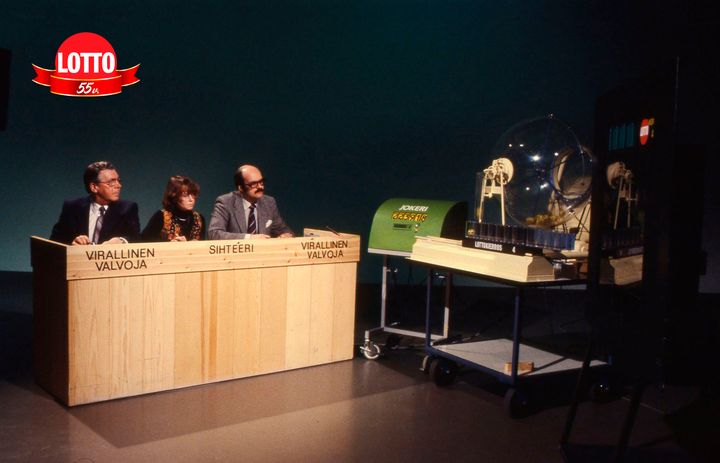 Kolme henkilöä istuu lottostudiossa vuonna 1981, seuraten arvontaa.