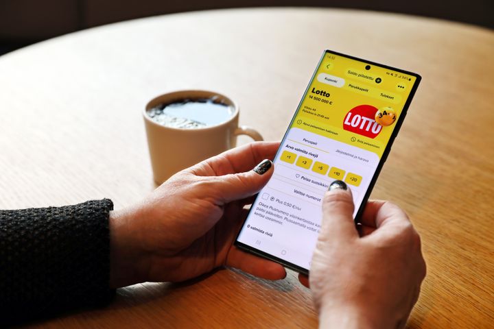Henkilö katsoo älypuhelimelta Lotto-sovellusta kahvikupin ääressä.
