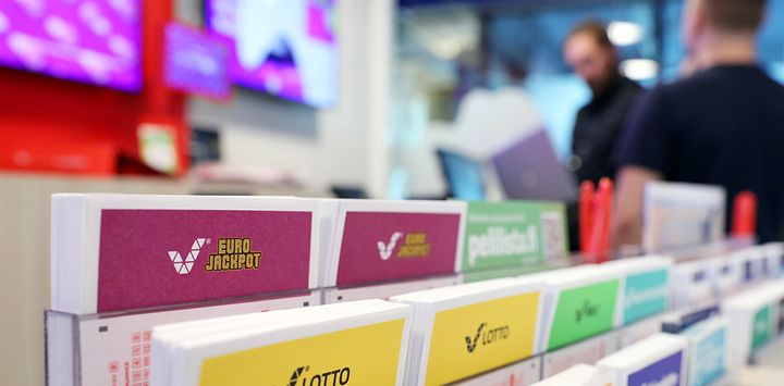 Kuvassa Eurojackpot-pelin kuponki telineessä muiden Veikkauksen pelien kuponkien kanssa. Taustalla ihmisiä.