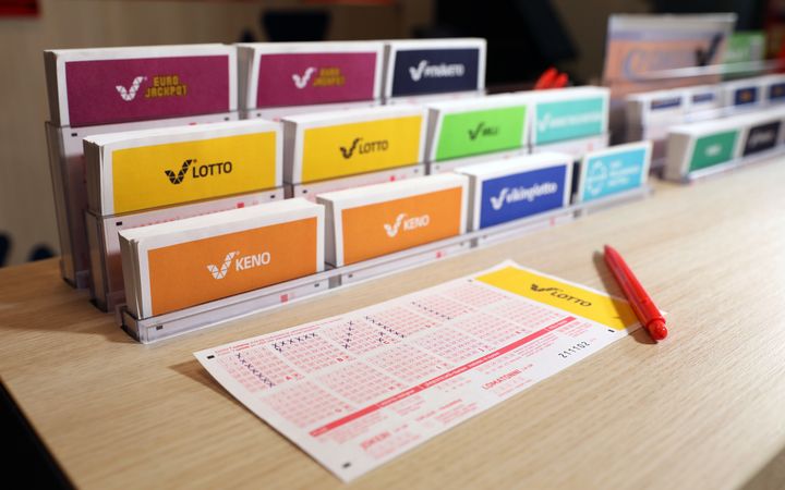 Lotton ja Keno-pelien kuponkeja telineessä pöydällä, etualalla täyttämättömiä lotto-kuponkeja ja punainen kynä.