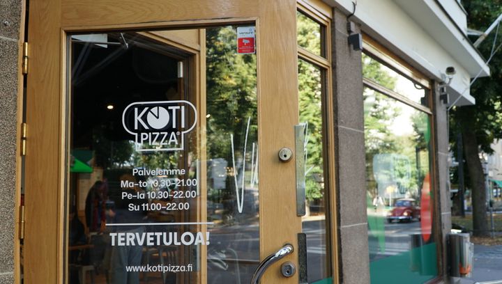 Kotipizza-ravintolan puukarminen lasiovi, jossa aukioloajat sekä Kotipizza-logo. Lasista heijastuu ulkona olevat puut.