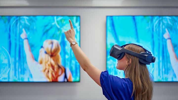Pitkätukkainen henkilö virtuaalilaseissa ja sinisessä paidassa osoittaa ylös vasemmalla kädellä, taustalla on kaksi suurta näyttöä.