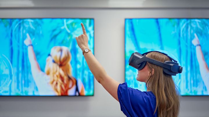 Pitkätukkainen henkilö virtuaalilaseissa ja sinisessä paidassa osoittaa ylös vasemmalla kädellä, taustalla on kaksi suurta näyttöä.