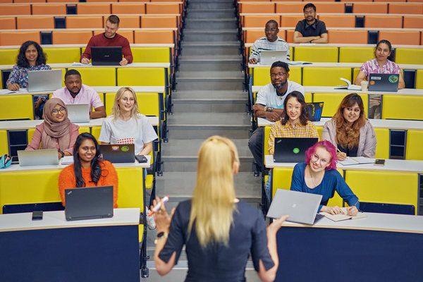 Luentosalissa opiskelijoita istumassa ja selin kameraan vaalea pitkähiuksinen opettaja.