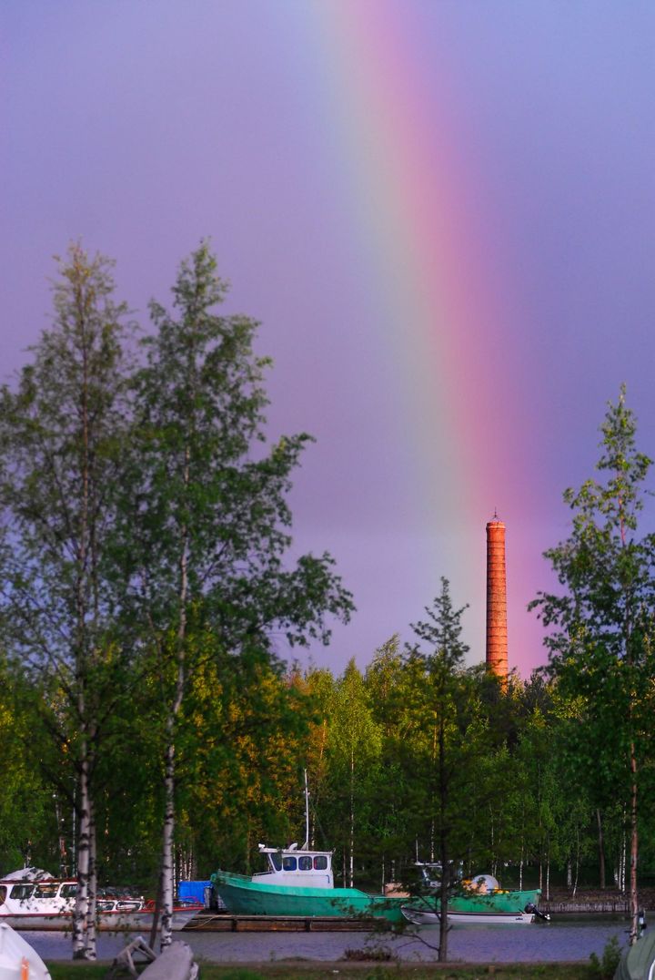 Puiden keskellä näkyy tehtaan punatiilinen piippu, johon osuu sateenkaari. Etualalla laiturissa kiinni veneitä.