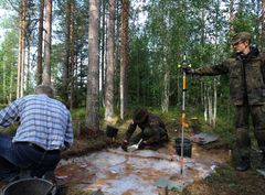 Kolme tutkijaa metsän keskellä olevan arkeologisen kaivauksen äärellä.