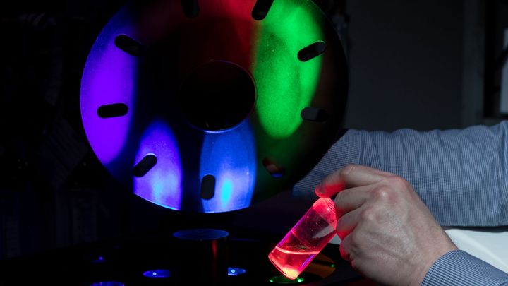 Metallisella kehällä heijastelee vihreää, punaista, sinistä ja violettia valoa, käsi pitelee pientä punaista nestettä sisältävää pulloa.