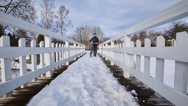Henkilö taluttaa polkupyörää lumisella sillalla, jossa valkoiset kaiteet.