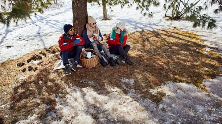 Kolme henkilöä istuu puun alla retkellä eväkori mukanaan. Puun alla ruskea nurmikko ja ympärillä vielä lunta.