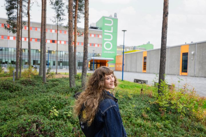 Nainen kävelee metsäisessä ympäristössä, taustalla näkyy Oulun yliopisto.