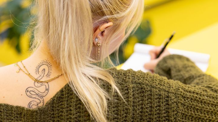 Henkilö, jolla on pitkä vaalea poninhäntä ja tatuointi niskassa, kirjoittaa muistiin vihkoon.