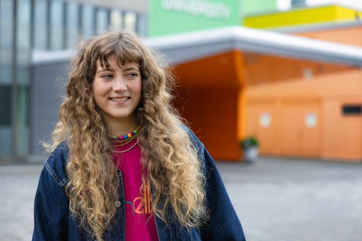 Nuori nainen seisoo ulkona hymyillen. Hänellä on pitkät kiharat hiukset ja värikäs paita. Taustalla on Oulun yliopiston pääsisäänkäynti.