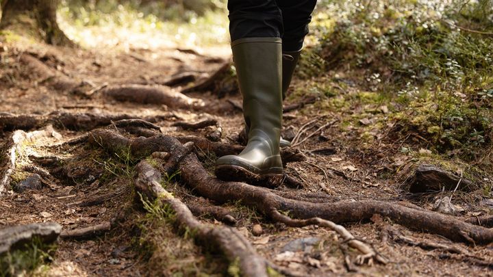 Henkilö kävelee metsäpolulla päällä vihreät kumisaappaat, polulla juuria ja lehtiä.
