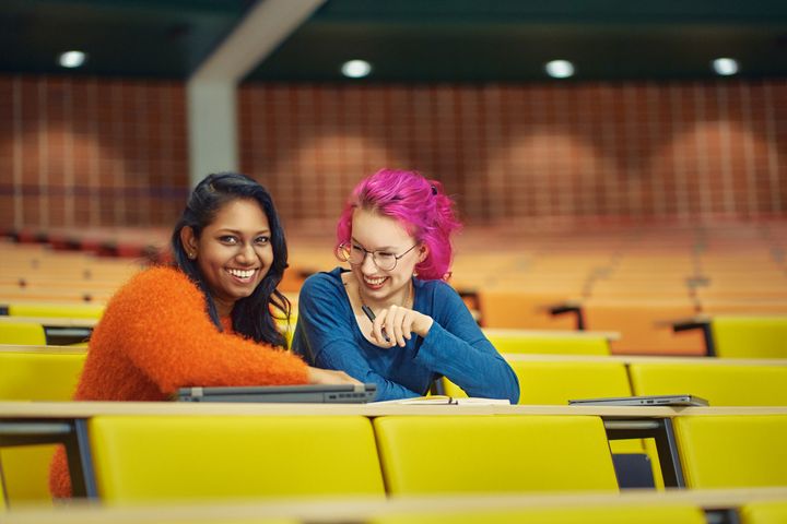 Kaksi hymyilevää henkilöä istuu värikkään luentosalin penkeillä ja katsoo tietokoneen näyttöä.