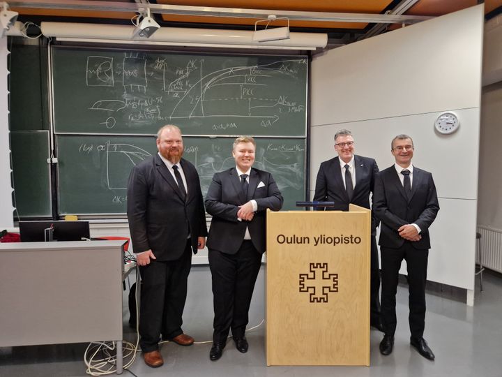 Kuva neljästä miehestä puvuissa seisomassa puhujakorokkeen takana Oulun yliopiston opetustilassa.
