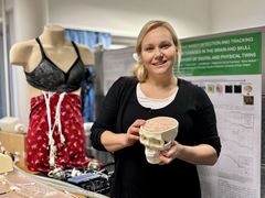 Alt-teksti: Henkilö laboratoriossa, jossa hän pitelee aivomallia. Taustalla on nukke, jolla on päällä monitorointiliivit.