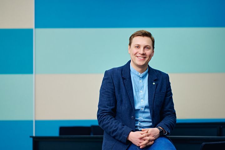 Kuvassa nuori mies sinisessä puvussa seisoo hymyilevänä. Taustalla seinä, jossa on sinisen sävyisiä raitoja.