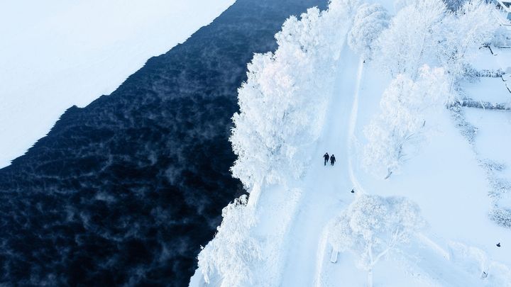 Talvinen ilmakuva lumisesta maisemasta, jossa kaksi henkilöä kävelee lumista polkua pitkin jäätyneen veden vieressä.