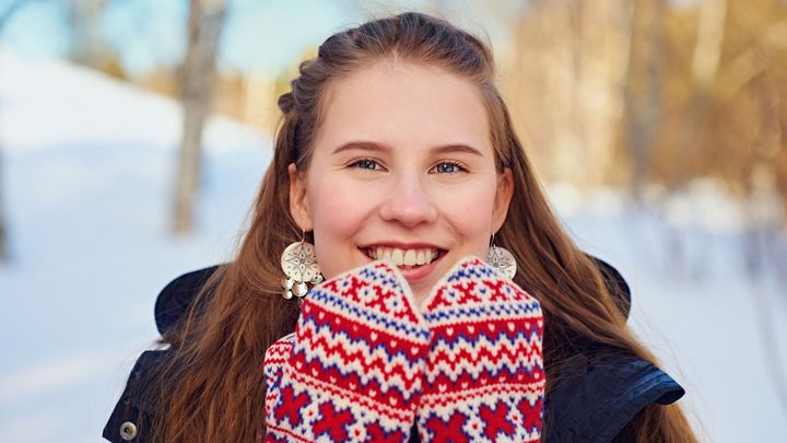 Nuori nainen hymyilee talvimaisemassa käsissään värikkäät kuviolliset lapaset.