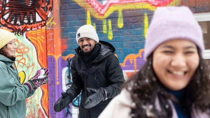 Kolme henkilöä ulkona talvivaatteissa, värikäs graffitiseinä taustalla.