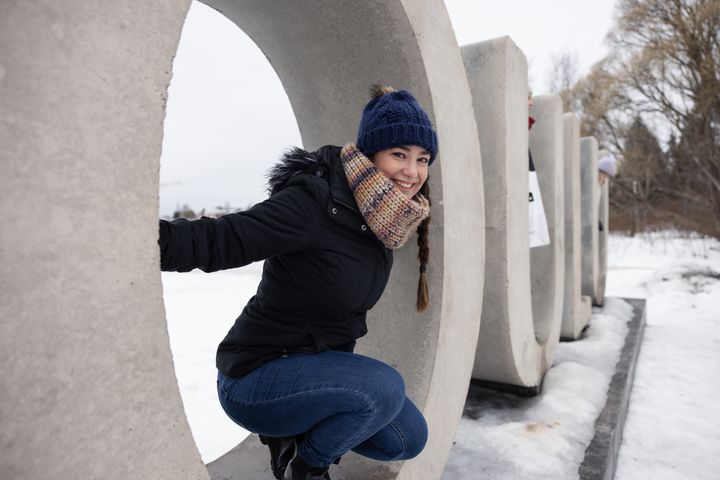 Nuori nainen hymyilee ja istuu suuren betonisen renkaan sisällä lumisessa ulkoilmassa. Betonikirjaimista muodostuu sana Oulu.
