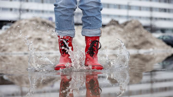 Punaisissa saappaissa oleva henkilö kävelee vesilätäkössä, roiskuttaen vettä ympärilleen kaupunkiympäristössä.