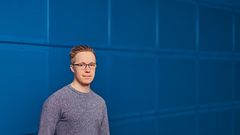 Nuori henkilö seisoo sinisen seinän edessä ja katsoo kameraan.