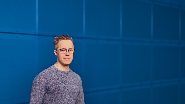 Nuori henkilö seisoo sinisen seinän edessä ja katsoo kameraan.