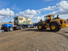 Etualalla kuormaaja liikuttaa maastokuvioitua konttia, taustalla näkyy kuorma-auto.