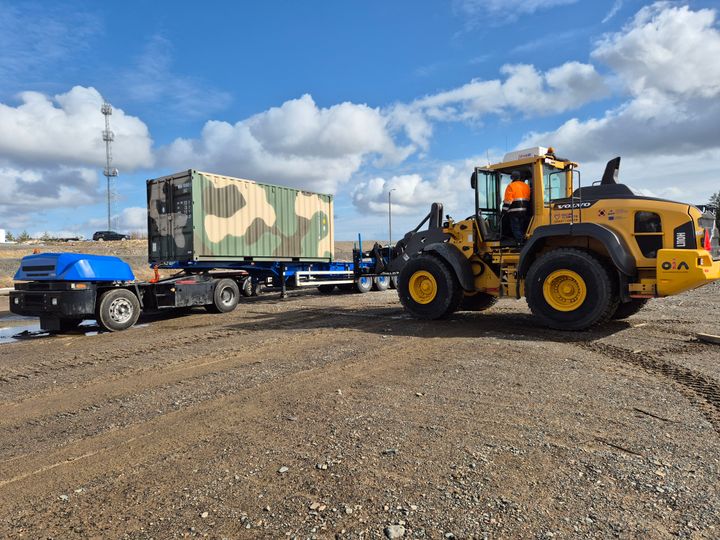 Etualalla kuormaaja liikuttaa maastokuvioitua konttia, taustalla näkyy kuorma-auto.