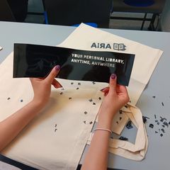 Henkilö pitää mustaa tarraa, jossa lukee: "Your personal library, anytime, anywhere". Taustalla kankainen kassi ja irtokirjaimia pöydällä.